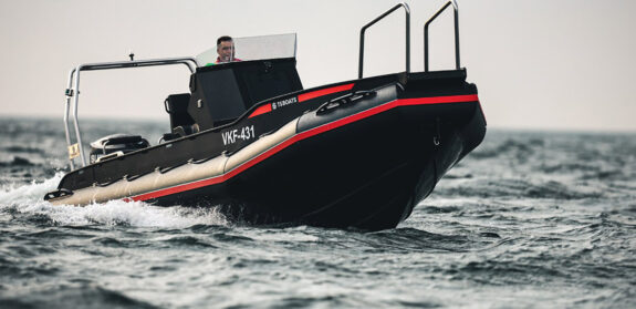 Side profile of a TS Boats craft boat model with a black and red hull, manned by a person in a casual stance, cruising on a grey, wavy sea.