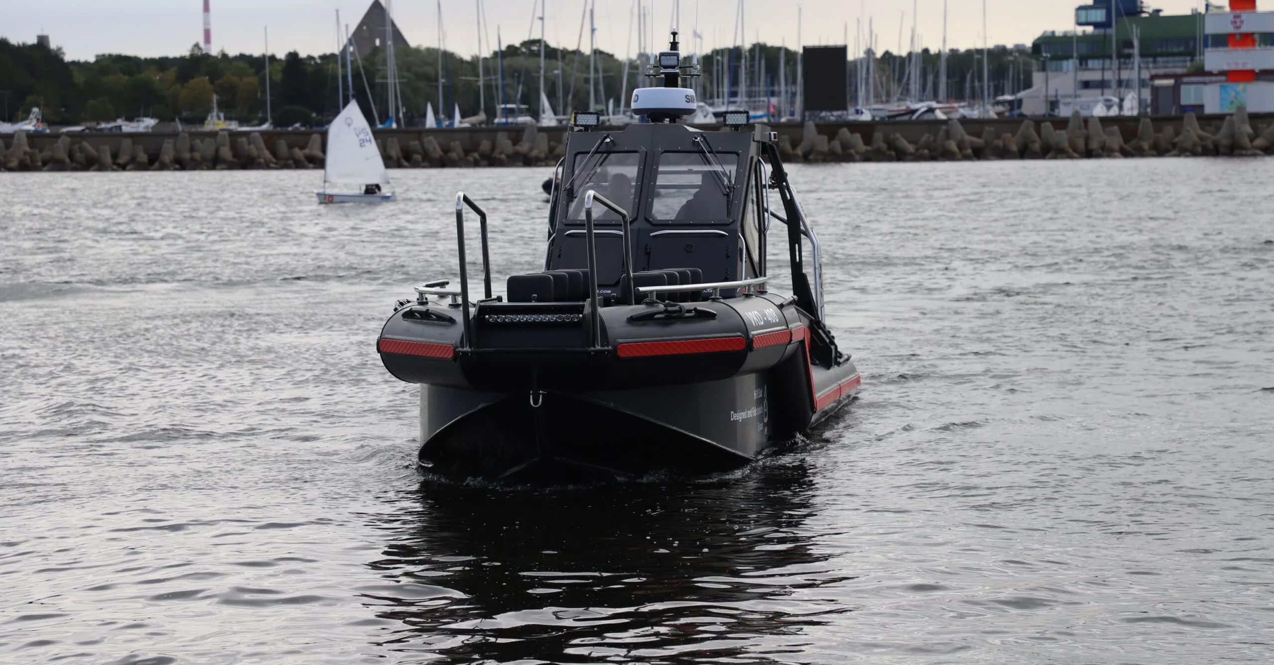 View of a TS Boats vessel on calm waters, featuring a cabin and dual outboard motors, with a marina and sailing boats in the background